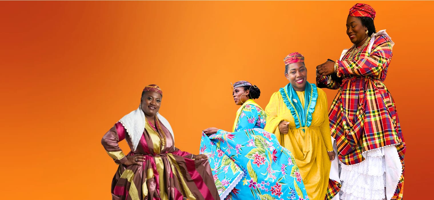 Four Women wearing different colour Wob Dwiyets, which is the national dress of Dominica. The first has on a striped wob dwiyet, the second has on a floral wob dwiyet, the third has on a gold wob dwiyet and the fourth is wearing a madras wob dwiyet, floral wob dwiyet and a striped wob dwiyet.